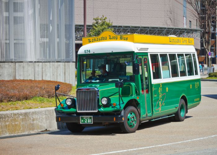 The Kanazawa Loop Bus on the road. It is bright green with a yellow sign, saying "Kanazawa Loop Bus" in a fancy font.